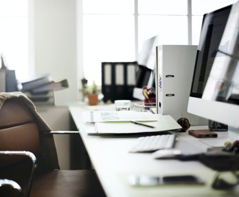 A desk and chair with jacket, papers, pen and desktop computer