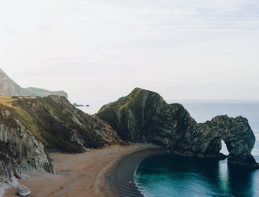 Durdle door, an arch landform, which is located at the far end of a small bay. The sea is calm and looks blue / green. The bay is surrounded by cliffs that are covered in vegetation.