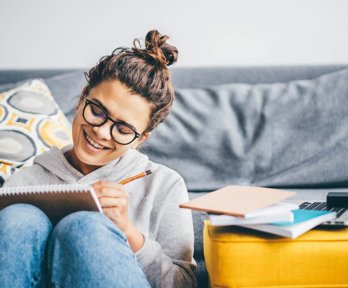 Woman writing in notebook, sitting in front of grey sofa, next to desk with laptop and papers