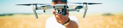 A man standing in yellow field of wheat, holding a silver drone out towards the camera. The drone obscures his face.