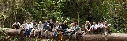 Group of people sitting on a large horizontal tree trunk surrounded by forest.