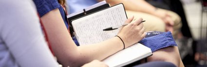 Woman writing in notebook, seated next to other people