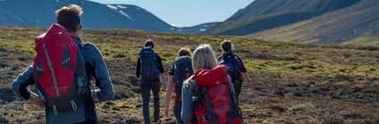 Five students wearing rucksacks viewed from behind walking through a grassy valley.