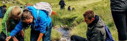 A group of students standing in a shallow upland stream, using a measuring tape to measure the width of the stream. In the background are green valley sides.