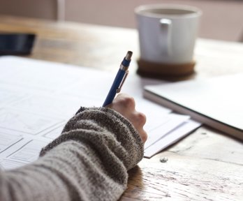 Person sat a table holding a pen and writing on a large piece of paper. On the table there is a mug on a coaster and a notebook.