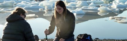 Two people sat beside a glacial lake taking measurements. They are surrounded by small transparent tubes and other measuring equipment.