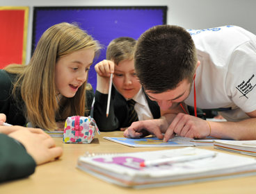 A young girl and boy sit at their desk in a classroom looking at a map with an adult geography ambassador.