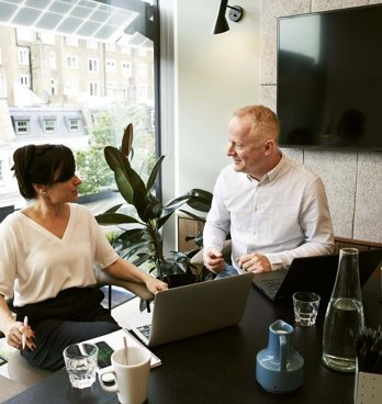 Two professionals having a conversation in an office with laptops in front of them