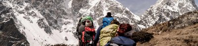 A group of people walk up mountainous terrain. They each carry large bags and walking equipment.