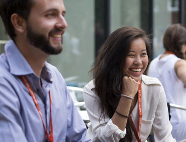 An Asian woman and white man sit next to each other smiling while wearing professional clothing and lanyards.