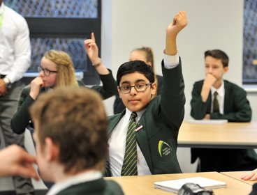 A young boy sits in his classroom wearing school uniform with his hand raised above his head.