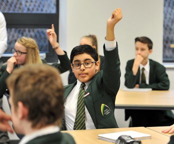 A young boy sits in his classroom wearing school uniform with his hand raised above his head.