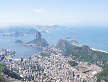 A view over Rio de Janeiro showing a sprawling city located in the valley between green wooded hills, finishing at the ocean