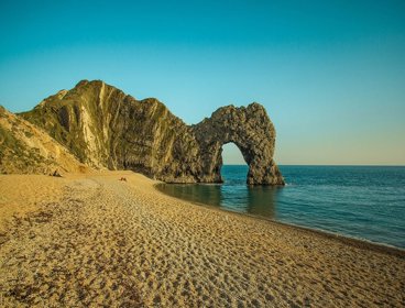 Durdle Door, an arch landscape formation, shows an arch in the rocks at the end of a small coastal sandy bay. The sun is low in the sky casting a soft light onto the rocks and the sea, which is very calm