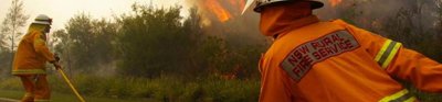 Two firemen in orange uniforms fight a fire by the side of the road. The flames are reaching high into the air and there is lots of smoke