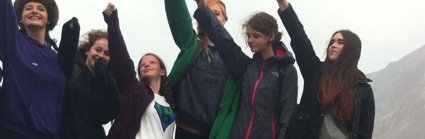 A group of young women standing on a rock in the outdoors with their arms reaching int the air, clearly enjoying geography