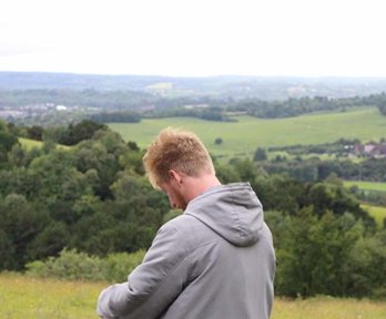 A male teacher standing in the countryside, writing on a clipboard. There is a lovely rural view behind him with green fields and trees.