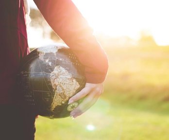 A person holding a small globe in their hand, down by their side. The sun is low in the sky and the light is bouncing off the top of the globe