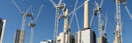 A city scene with skyscrapers and cranes to illustrate the amount of building work going on. In the foreground are colourful adverts on a boarded wooden wall around the building site.