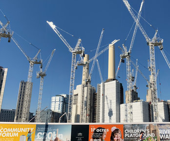 A city scene with skyscrapers and cranes to illustrate the amount of building work going on. In the foreground are colourful adverts on a boarded wooden wall around the building site.