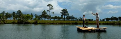 Two people standing on wooden raft on a lake, with a grassy bank and trees in the background. 