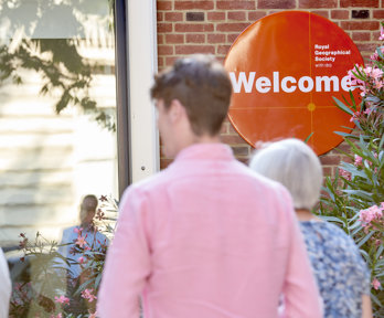 People walk into the RGS at Exhibition Road. An orange welcome sign is focused in on.