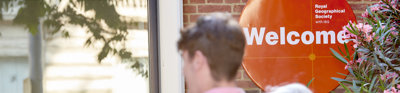 People walk into the RGS at Exhibition Road. An orange welcome sign is focused in on.