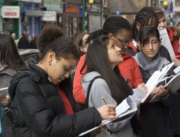 A group of female school children, holding clipboards and looking at buildings on a high street