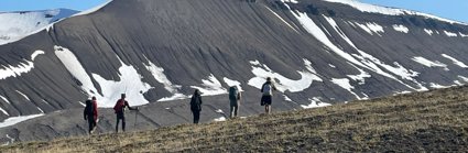 Five people walking in a line through a mountainous landscape. The mountain slope is largely bare with patches of snow remaining. 