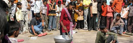 A large group of people, with children at the front, gathered looking towards a women standing in the middle with a metal bowl placed on a metal barrel. 