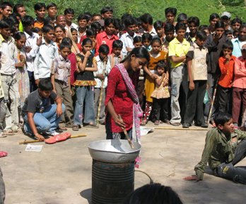 A large group of people, with children at the front, gathered looking towards a women standing in the middle with a metal bowl placed on a metal barrel. 