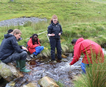 A group of teachers collected by a small fast flowing stream, three are sitting on the banks and two are standing in the stream. One is measuring the depth of the stream with a stick.