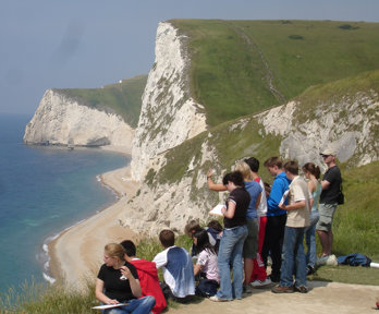 A group of students are sitting near the edge of a cliff, with a view down onto a sandy beach with white chalk cliffs below them.