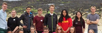 A group of older students lined up on a beach. In the background you can see a rocky beach with seaweed, and then the sea.