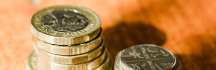 Two piles of english coins, pound coins and two pound coins to the left hand side and fifty pence, twenty pence and five pence to the right hand side, They are placed on a wooden table.