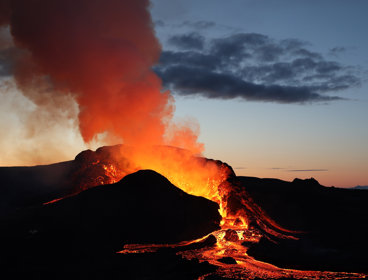 An erupting volcano, with orange and yellow lava running down the hill away from it. Smoke coming from it is tinged orange by the heat. 