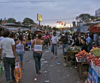 People walking through an outdoor market with fresh fruit and vegetables displayed in crates on market stalls. 