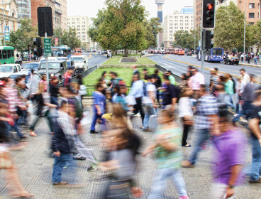 A busy street crossing with lots of people. The people are blurred to suggest that they are moving quickly. In the background is a city, cars and a tree