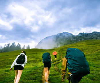 Four people with large backpacks are walking up a green hill towards a rocky outcrop. The clouds are low and are forming a mist at the top of the hill towards where they are heading