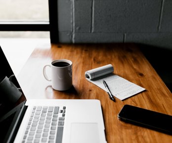 A laptop, coffee mug containing coffee, notebook, pen and mobile phone on a desk