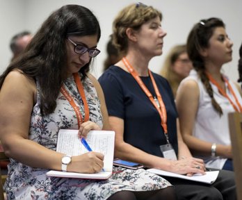 A woman writing in a notebook while listening to a conference presentation, seated next to two other conference delegates