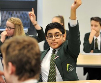 A young male student sitting at a desk with his hand up, ready to answer a question