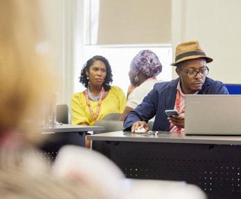 A man wearing a hat and glasses using a laptop, in the background two women having a conversation