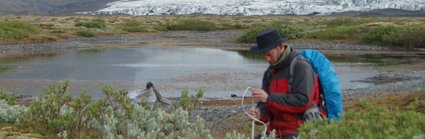 Person setting up a measuring device in front of a glacier and lake. The person is wearing a hate and rucksack. 