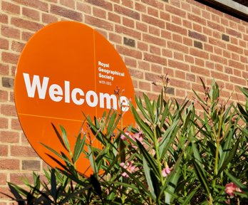 A circular orange sign reading Welcome, next to a green plant with pink flowers