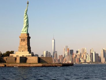 A view from the river of New York City, showing the Statue of Liberty and a skyscraper horizon. The sun is low in the sky so the light is soft