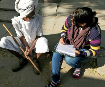 Two people sat on the a concrete pavement conversing. The person on the right is holding a clipboard and pen and making notes.