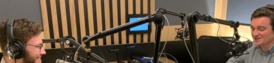 Two men sitting across from eachother at a wooden desk, both wearing headphones. Both have microphones and sound recording equipment around them and one is interviewing the other