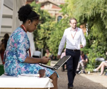 A seated woman next to a marquee in a garden, using a mobile phone and laptop