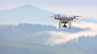 A grey and orange drone flying in the sky, a blurry background of forested hills can be seen